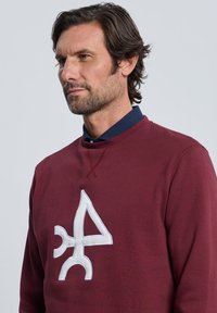 Sudadera burdeos con cuello redondo, que presenta un gran logo abstracto blanco en el pecho. Fabricada con un tejido suave y texturizado.