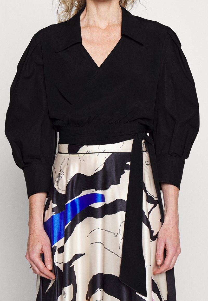 Femme portant un chemisier noir cache-cœur à manches bouffantes et une jupe taille haute avec des motifs abstraits noirs et bleus sur un tissu crème.