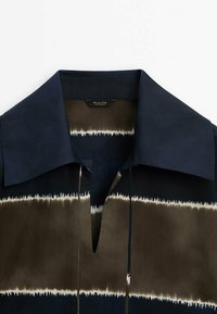 Chemise boutonnée avec col bleu marine, présentant des rayures horizontales en brun foncé et beige clair, avec une encolure fendue et une texture lisse.