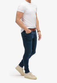 Camiseta blanca de manga corta combinada con pantalones ajustados de mezclilla azul oscuro y zapatillas beige. Los pantalones tienen bolsillos y un cinturón marrón con accesorios metálicos.