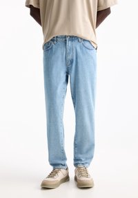 Jeans de mezclilla azul claro con un corte relajado, diseño de cinco bolsillos y una textura ligeramente desgastada, combinados con zapatillas beige y una camiseta suelta beige.