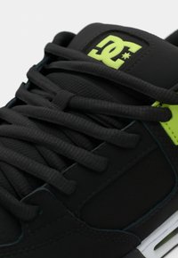 Schwarze Sneakers mit strukturierter Oberfläche, flachen schwarzen Schnüren, einem auffälligen gelben DC-Logo und weißen Sohlenakzenten.