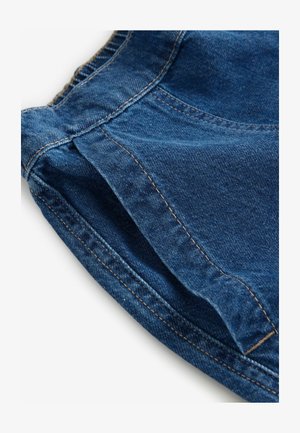 Tummansininen farkkukangas, jossa näkyvät ompeleet. Sisältää laskoksia ja kevyen kaaren vyötäröllä, korostaen jämäkkää muotoilua.