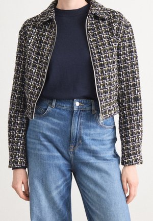 Person iført en mønstret tweedjakke med lynlås over en navyblå sweater, kombineret med blå denimjeans med høj talje mod en neutral baggrund.