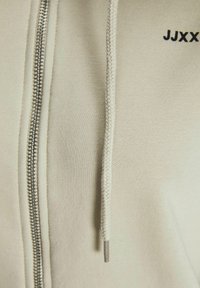 Sweat à capuche zippé beige avec une texture lisse, doté d'un zip métallique et d'un cordon de serrage tressé. Le nom de la marque "JJXX" est brodé en noir.