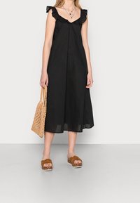 Robe noire sans manches avec épaules à volants, longueur midi, portée avec des sandales à plateforme marron nouées et un sac à main tissé beige.