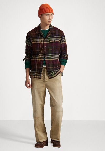 Flanellen shirt met een veelkleurig ruitpatroon, gecombineerd met een groene trui, beige broek en bruine schoenen. Oranje gebreide muts op het hoofd.