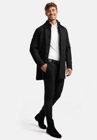 Sort trenchcoat med knapper, hvid polo-shirt, sorte slim-fit bukser og sorte ruskind ankelstøvler. Model står med hænderne i lommerne.