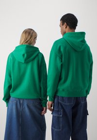 Sweatshirt à capuche verte avec une coupe décontractée ; fabriqué en tissu doux, avec des poignets et un ourlet côtelés. Présenté sur deux individus de dos.