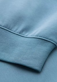 Jasnoniebieski materiał bluzy z teksturowanym splotem i gładkim ściągaczem. Posiada dopasowany krój z ribbowanym dołem i miękką powierzchnią.