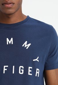 Navyblaues Baumwoll-T-Shirt mit weißen Buchstaben, die in einem kreisförmigen Design angeordnet sind und "TOMMY" und "HIGER" schreiben. Es hat kurze Ärmel und einen Rundhalsausschnitt.