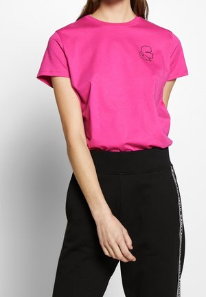 Mujer con camiseta de manga corta de color rosa brillante con un pequeño diseño de perfil en contorno negro en el pecho, combinada con pantalones negros de cintura alta.