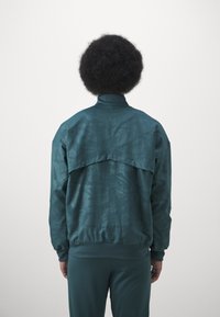 Giacca turchese con colletto alto, caratterizzata da un motivo testurizzato. La parte posteriore presenta un elemento di design distintivo che aggiunge interesse visivo.