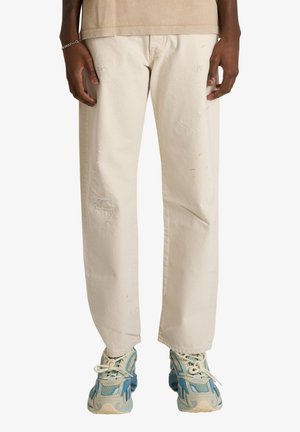 Persona che indossa pantaloni dritti color ecrù usurati e scarpe da ginnastica blu e bianche, in piedi con le mani rilassate ai fianchi.