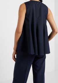 Mörkblå, ärmlös topp med avslappnad passform, har en rundad halsringning och dragkedja bak, tillverkad av ett mjukt tyg med en svävande design.