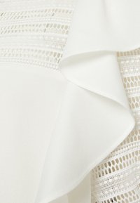 Blusa bianca con un tessuto morbido e testurizzato, caratterizzata da volant a strati. Decorata con inserti in pizzo e una finitura trasparente, creando un contrasto visivo.