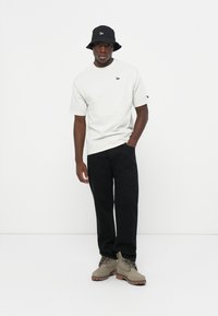 Light gray short-sleeve t-shirt with small black logo, black pants, and a black bucket hat, paired with gray boots featuring brown rubber soles.