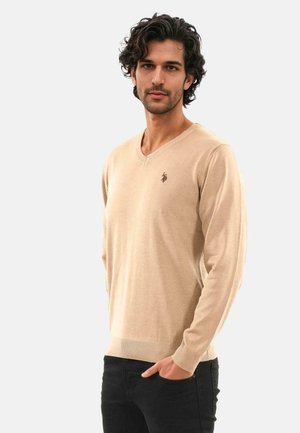 Beige v-neck sweater in soft knit material with ribbed cuffs and hem; features a small brown logo on the chest. Paired with black pants.