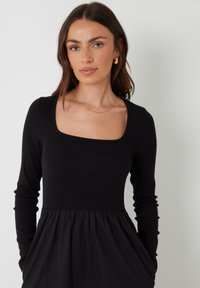 Vestido negro de manga larga con torso acanalado y escote cuadrado, que fluye hacia una falda fruncida. Cuenta con bolsillos laterales y una textura suave.