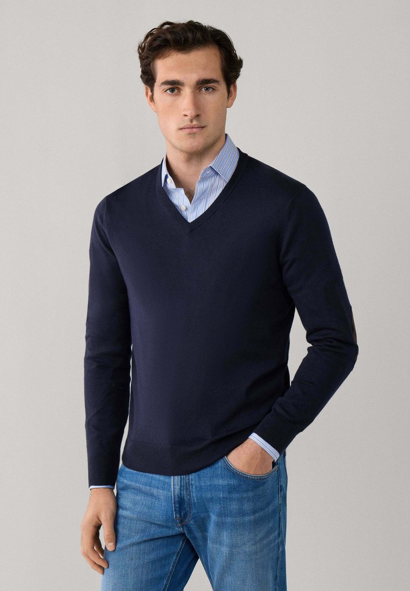 Giovane uomo con maglione a scollo a V blu navy sopra camicia a righe blu, con jeans, in piedi con una mano in tasca contro uno sfondo semplice.