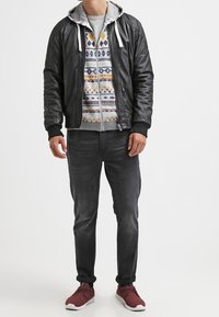 Chaqueta bomber de cuero negro sobre una sudadera con patrón, jeans grises y zapatillas deportivas rojas. Capucha con cordones visibles.