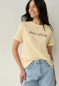Kollane lühikeste varrukatega T-särk, millel on üles keeratud varrukad, ja millel on pruun kiri "Hey chick". Koos heleda sinise teksapüksega. Pehme puuvillane tekstuur.