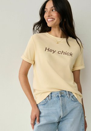 Geel T-shirt met korte mouwen en opgerolde mouwen, voorzien van de tekst "Hey chick" in bruin. Gepaard met lichtblauwe jeans. Zachte katoenen textuur.