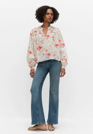 Vrouw staand, gekleed in een bloes met lange mouwen met bloemmotief, blauwe spijkerbroek en gouden sandalen met bandjes, tegen een effen lichte achtergrond.