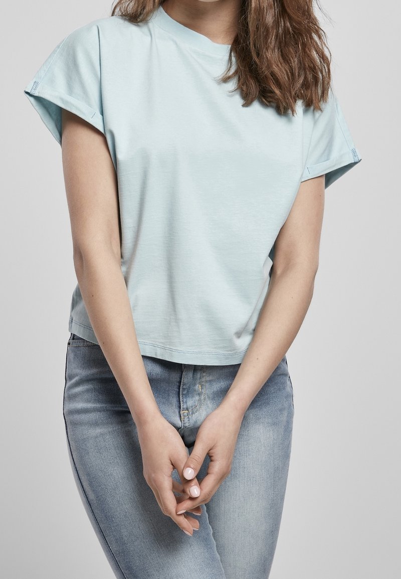 Femme portant une chemise à manches courtes bleu clair et un jean délavé clair, debout avec les mains légèrement jointes devant elle.
