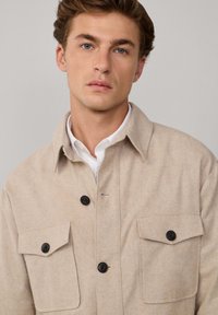Chaqueta de mezcla de lana beige con cierres de botón, dos bolsillos en el pecho y un collar, superpuesta sobre una camisa blanca. Textura suave, diseño casual.