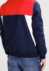 Chaqueta azul marino con acentos rojos y blancos, con un estilo bomber, puños elásticos y una textura suave y brillante.