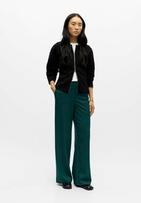Black zippered jacket, white shirt, green wide-leg trousers, and black shoes. Smooth fabric with a relaxed fit. Simple design and neutral colors.