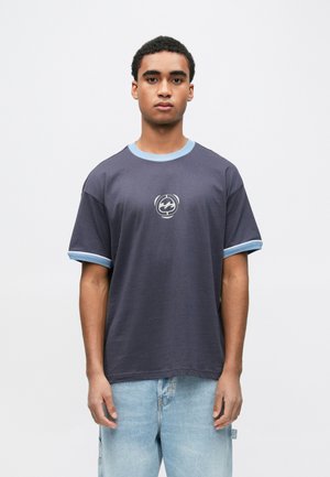 Young man wearing a navy blue t-shirt with light blue trim and a circular logo, paired with light blue denim jeans against a white background.