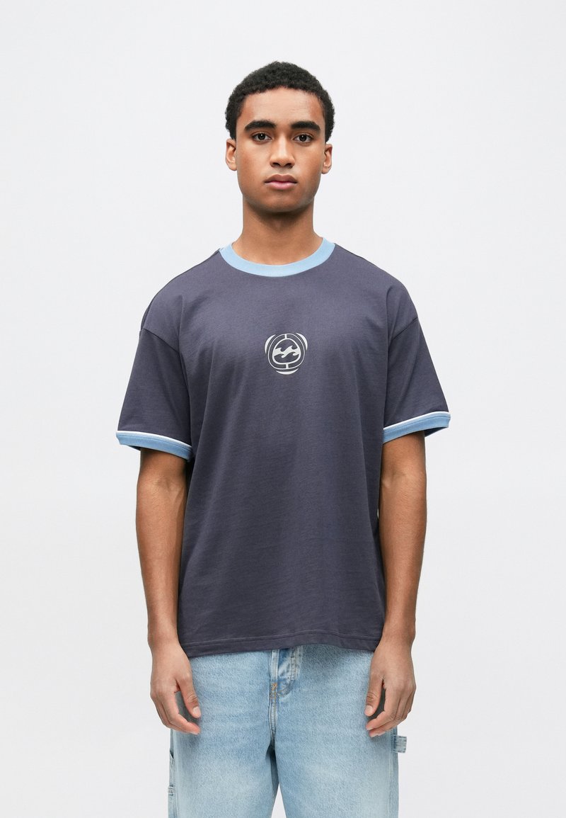 Νεαρός άνδρας φοράει μπλε μπλουζάκι με ανοιχτό μπλε περιθώριο και κυκλικό λογότυπο, σε συνδυασμό με ανοιχτό μπλε τζιν τζιν, πάνω σε λευκό φόντο.