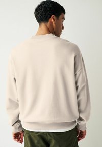Sweatshirt beige clair avec un col rond et des poignets côtelés, offrant une coupe décontractée et une texture douce. Porté sur un pantalon vert olive.