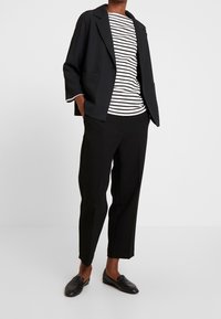 Blazer noir non structuré avec deux poches avant, porté sur un haut rayé blanc et noir. Pantalon noir fuselé et mocassins en cuir noir complètent la tenue.