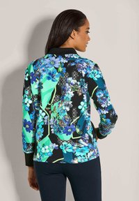 Frau in einer langärmeligen blumigen Jacke mit blauen, grünen und schwarzen geometrischen Mustern, von hinten gesehen.