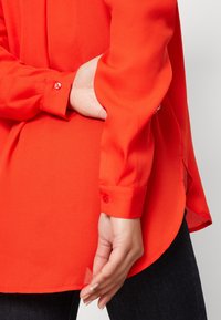 Blouse rouge à manches longues avec une texture lisse, poignets boutonnés et ourlet arrondi. Une main repose sur le côté, en contraste avec un denim foncé.