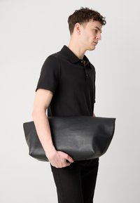 Čierna kožená taška cez plece s hladkým povrchom a zaobleným tvarom, držaná jednou rukou modelom v čiernej polo košeli.