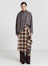 Maglione grigio texturizzato con bottoni, indossato sopra una gonna a quadri decorata con tonalità beige e marroni. Abbinato a pantaloni marroni e scarpe nere.