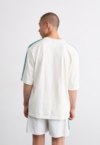 Cămașă albă cu mâneci scurte, cu dungi verzi pe laterale, croială lejeră și guler rotund, asortată cu pantaloni șort albi.