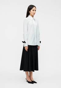 Λευκό πουκάμισο με μακριά μανίκια και μαύρες φιόγκους ως διακοσμητικά στοιχεία, συνδυασμένο με μαύρη, ροή μιντί φούστα και μαύρα παπούτσια με τακούνι. Απλός, δομημένος σχεδιασμός.