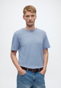 Šviesiai mėlynas medvilninis marškinėlis su trumpomis rankovėmis ir apvalia iškirpte, derinamas su laisvo silueto išblukusiais mėlynais džinsais ir ruda diržu.