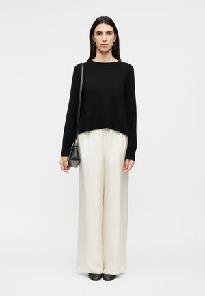 Sieviete ar gariem tumšiem matiem, valkājot melnu džemperi, platus baltus bikses, melnus apavus, zelta riņķa auskarus un nēsājot nelielu melnu plecu somu.