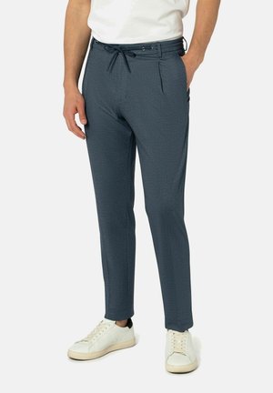 Mann in marineblauen Jogginghosen und weißen Sneakers, der mit einer Hand in der Tasche steht und die andere entspannt an der Seite hat.