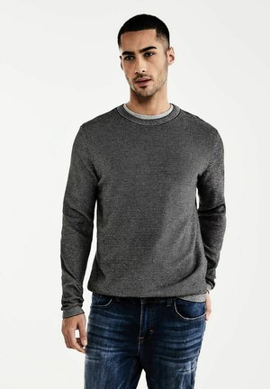 Maglione a maniche lunghe a righe nere e grigie con scollatura tonda, abbinato a jeans blu. Il tessuto ha una consistenza liscia.