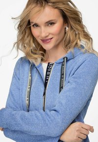 Blå zip-up hoodie med texturerad yta, med svarta dragsnören och logotypdetaljer. Känguruficka synlig framtill.