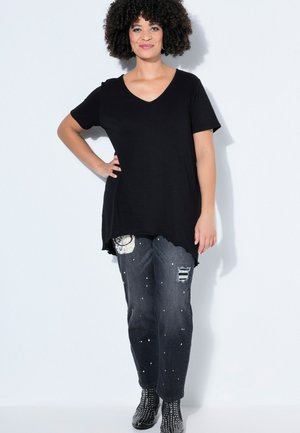 Frau mit lockigem Haar, die eine schwarze V-Ausschnitt-Tunika, abgenutzte graue Jeans mit weißen Sprenkeln und schwarze Nieten-Stiefeletten trägt und vor einer weißen Wand steht.