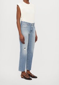 Jean en denim bleu clair avec une coupe décontractée, présentant des détails usés. Associé à un haut moulant en tricot crème et des chaussures plates noires.