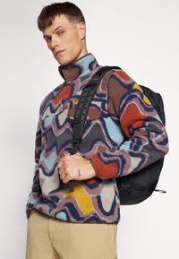 Fleece-paitsi, jossa on monivärinen aalto-kuvio, korkea kaulus ja ruskea rintatasku. Malli kantaa mustaa reppua.